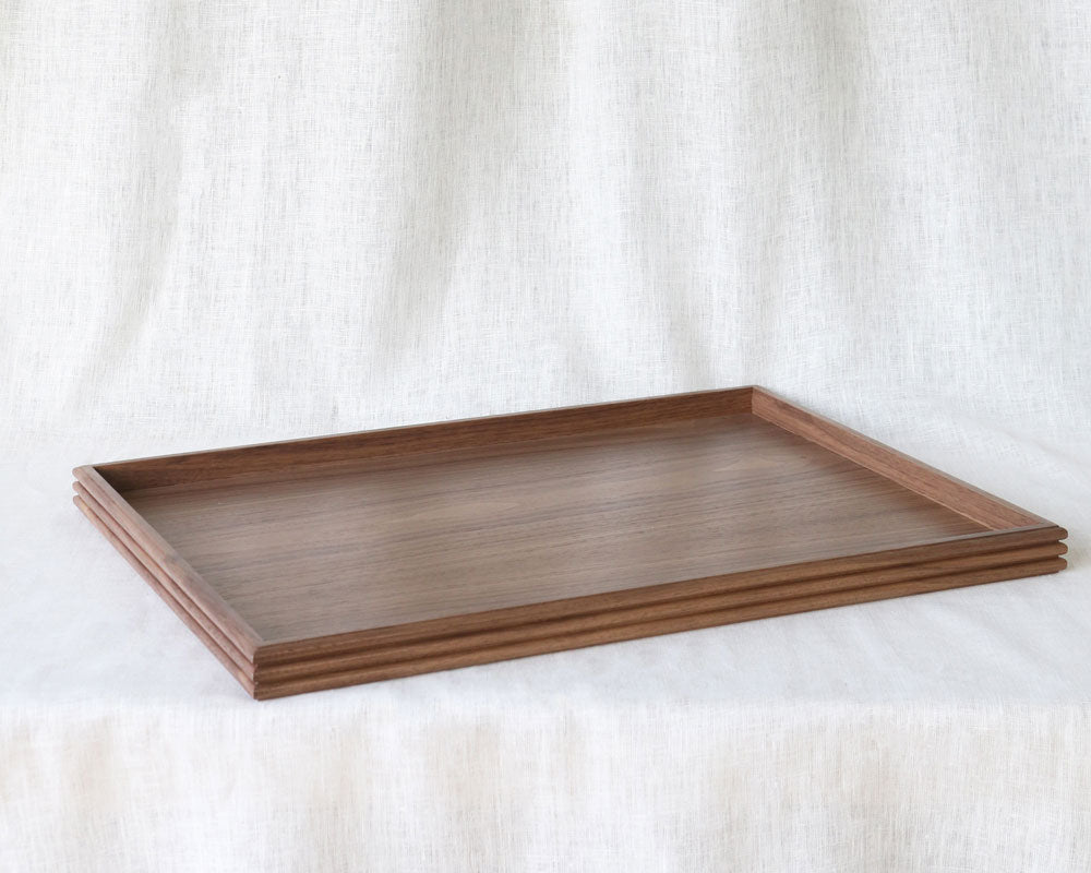 Angled view of an empty rectangular walnut wooden tray, showcasing its smooth surface and reeded edge detail, placed on a soft white linen background.