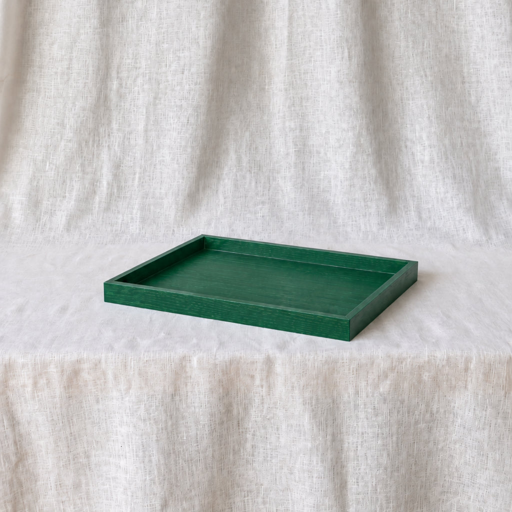 A square dark green tray with a raised rim, placed on a white textured fabric surface against a matching draped fabric backdrop.