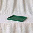A square dark green tray with a raised rim, placed on a white textured fabric surface against a matching draped fabric backdrop.