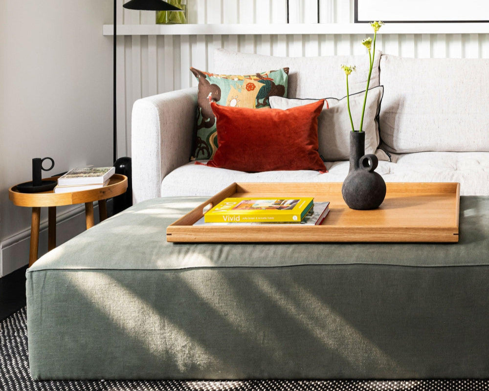 Large wooden ottoman tray placed on a green footstool in a cozy living room, featuring a yellow book and a sculptural black vase.